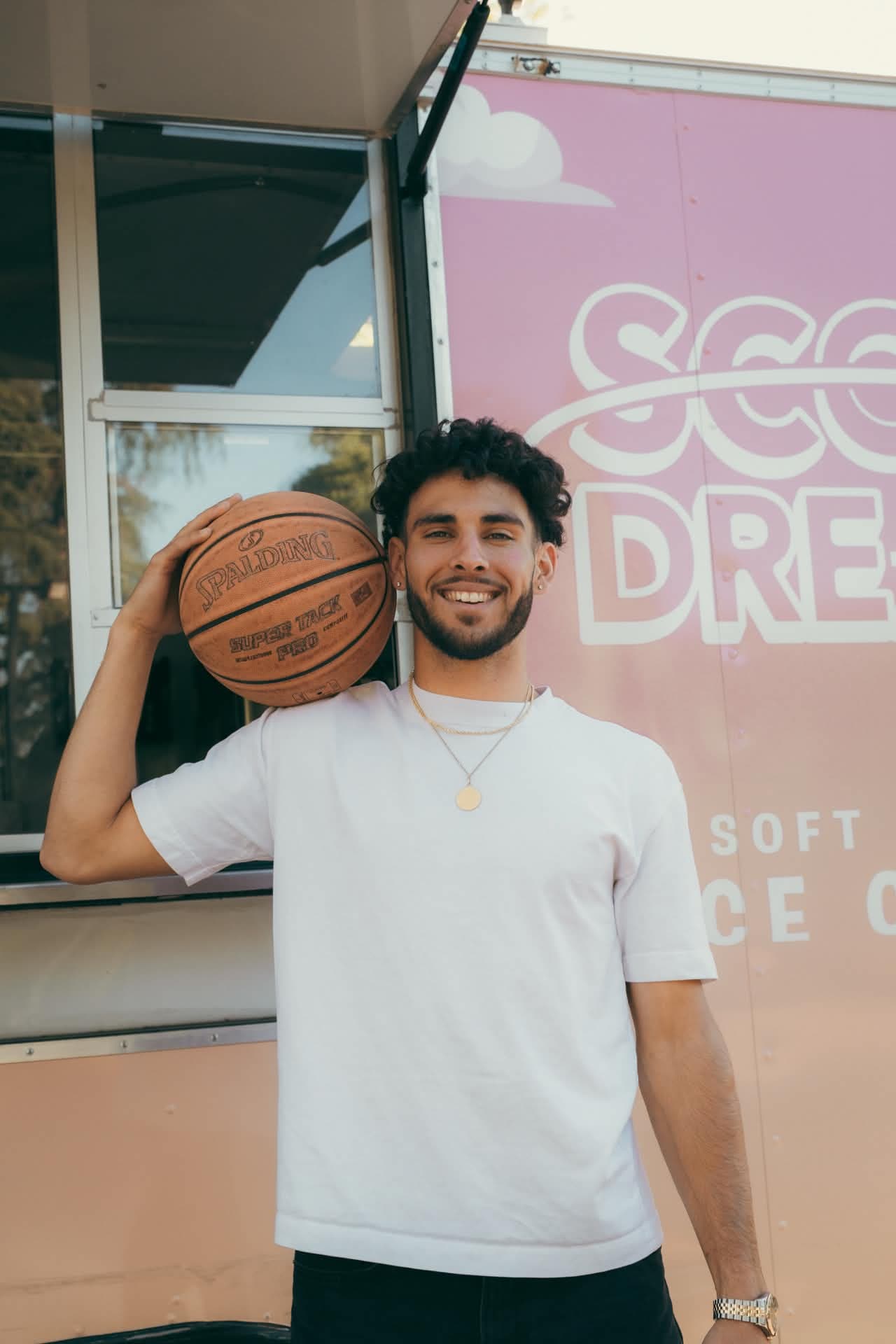 Devan Minouei smiling with a Spalding basketball over his shoulder in front of the Scoop Dreams trailer