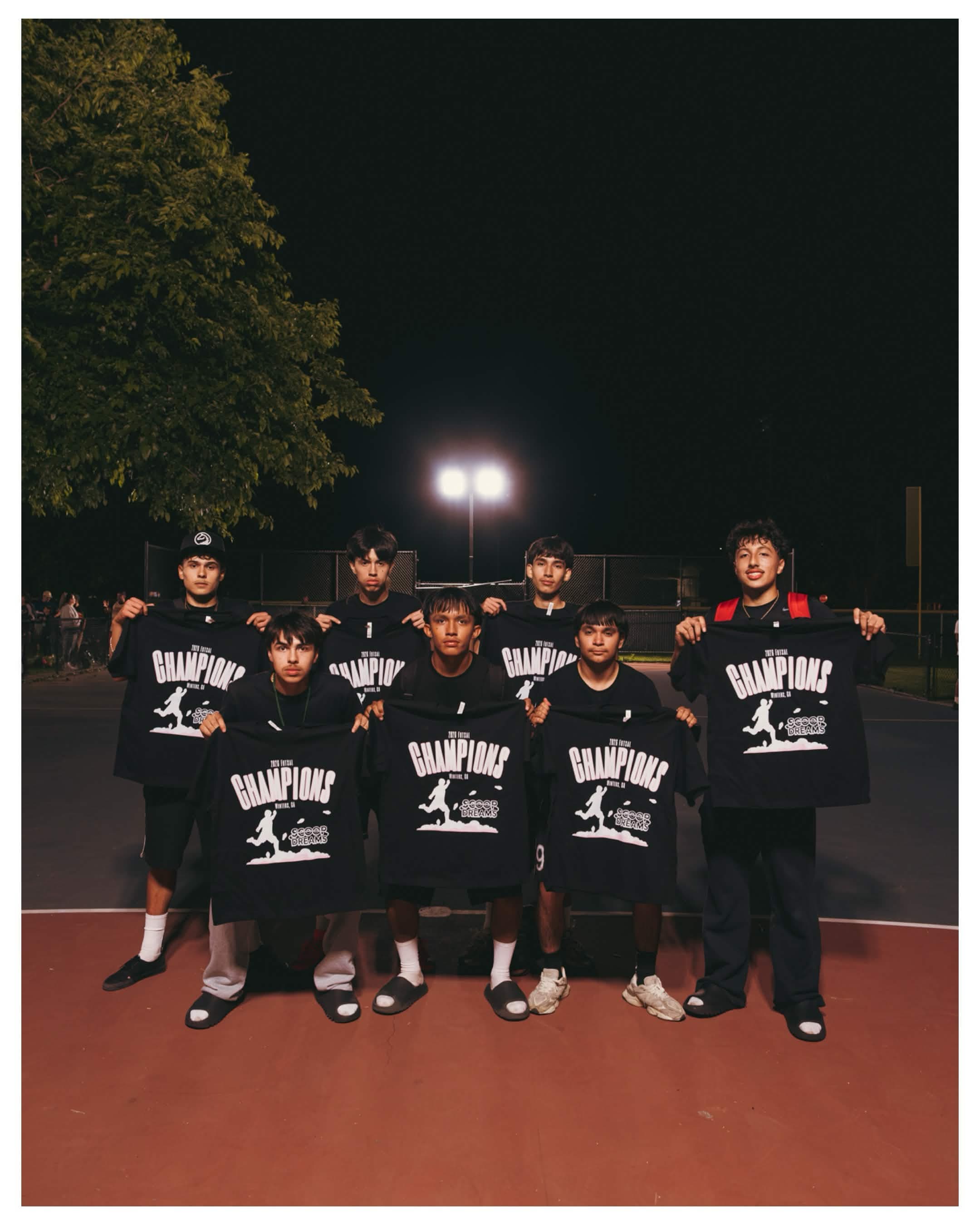 Championship team holding their Scoop Dreams 3-on-3 tournament shirts on the court at night